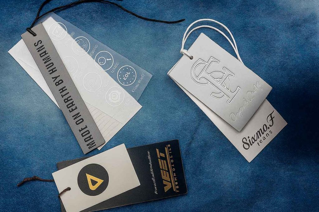 Etiquetas colgantes de doble cartón sobre fondo denim, con diseños personalizados y acabados de alta gama.
