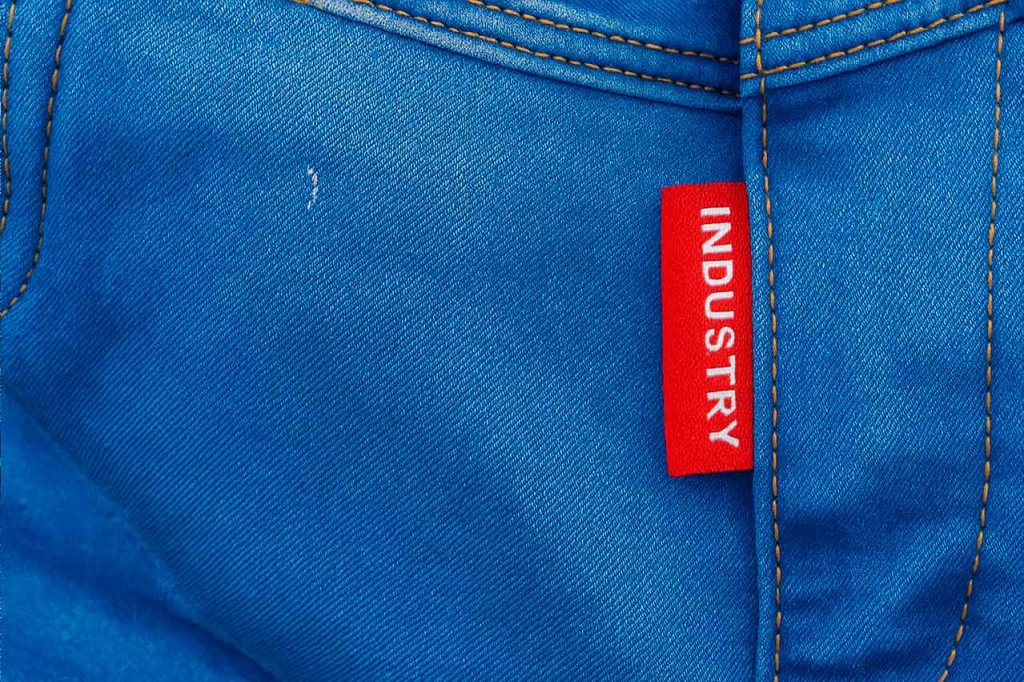 Marquilla punto tejida color rojo con texto INDUSTRY, cosida en el costado del bolsillo de un pantalón jean azul. Etiqueta tejida visible y personalizada.