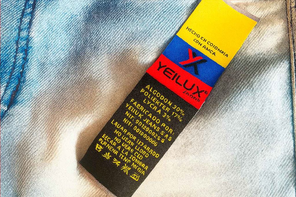 Marquilla tejida con símbolos e instrucciones de lavado de la marca YEILUX, fabricada en Colombia. Incluye composición textil y datos del fabricante.