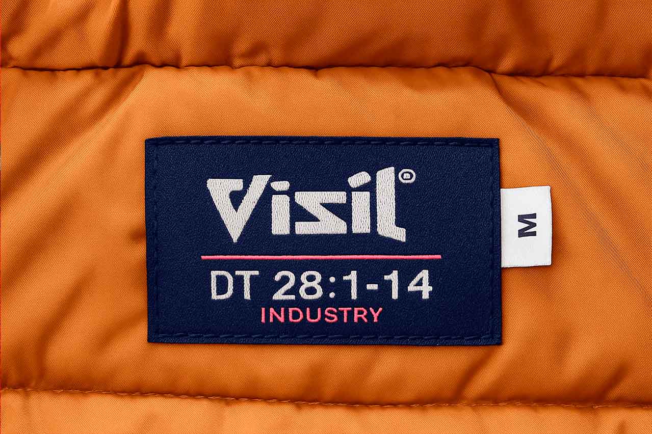 Etiqueta tejida de talla M personalizada de la marca VISIL, cosida en chaqueta color mostaza. Marquilla tipo cogotera resistente, con texto bordado y acabado profesional.